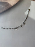 A silver chain bracelet with small dangling charms, photographed on a light gray surface with a soft shadow.